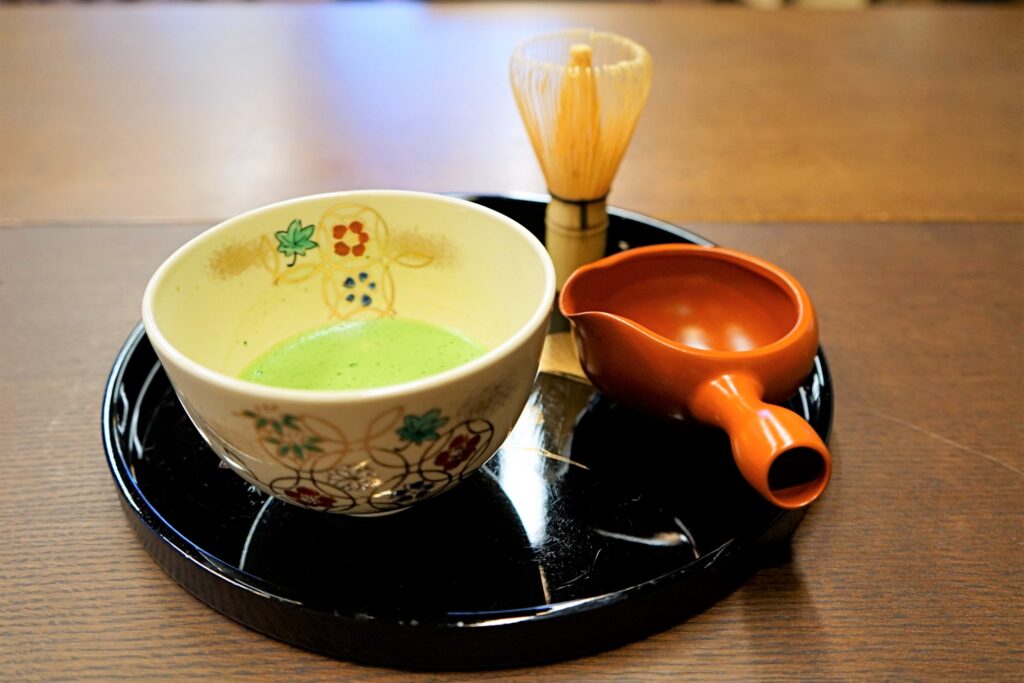 A matcha whisk, a bowl of matcha, and a red ladle on a plate